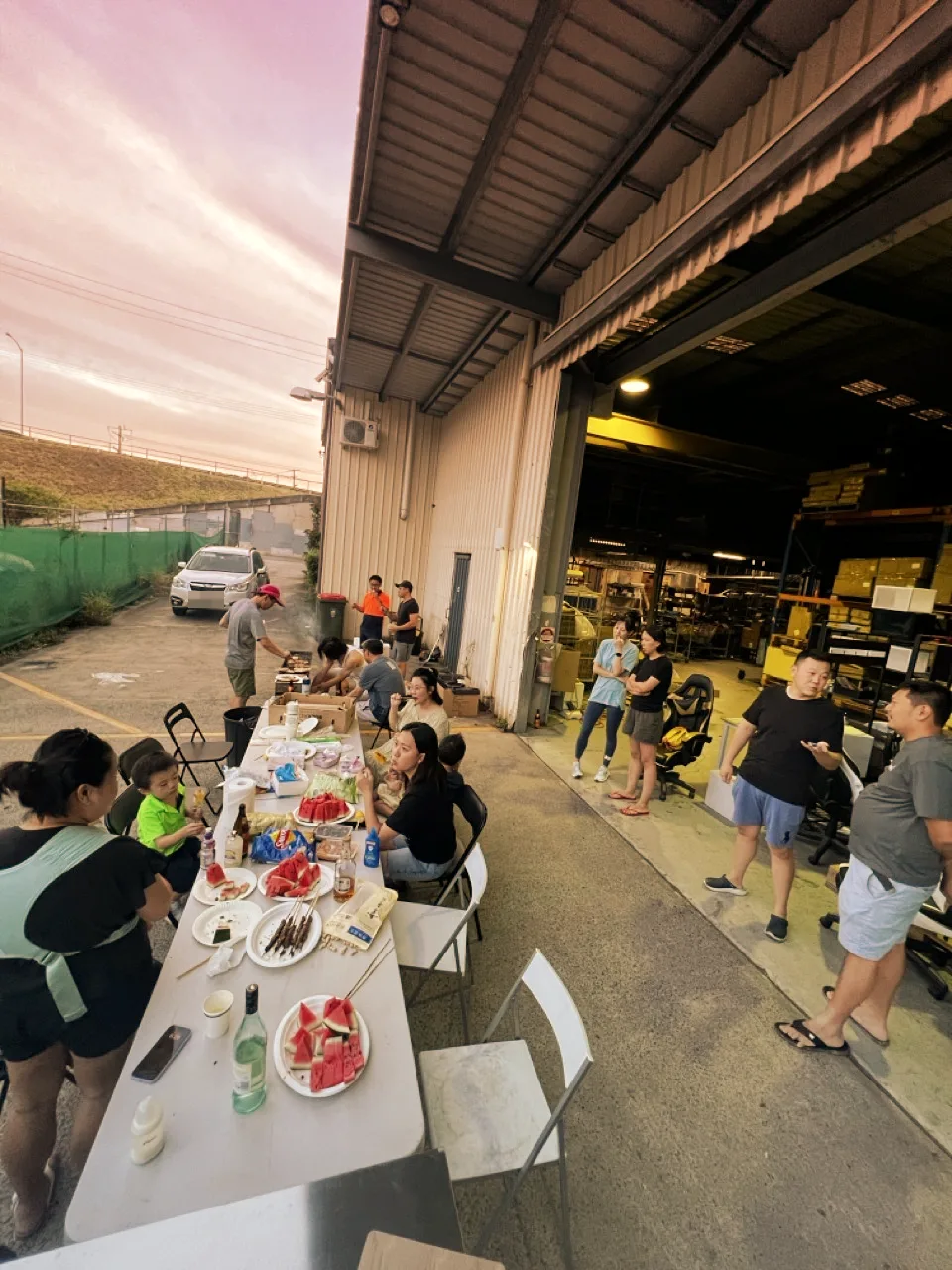 Sunset BBQ outside the open Canetoad Equipment warehouse: a long table with watermelon and skewers, people chatting by the roller door